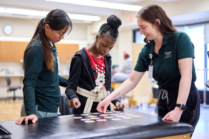Two student health-care providers stand with a patient (center) working on balance and motor skills.
