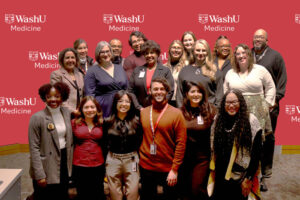Several individuals stand and kneel as a group in front of a red WashU Medicine backdrop at the Drum Major Awards ceremony.
