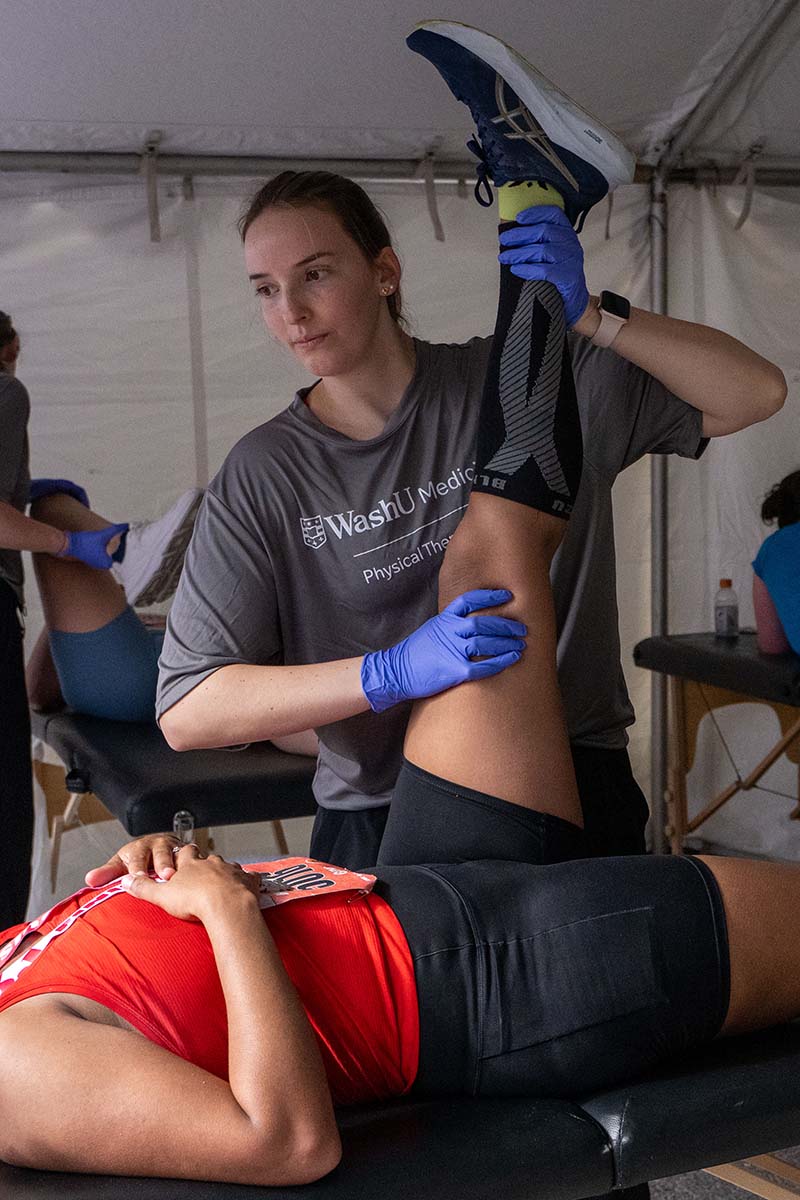 Race participant lays on bench while woman holds his left leg up in the air, trying to get a good stretch