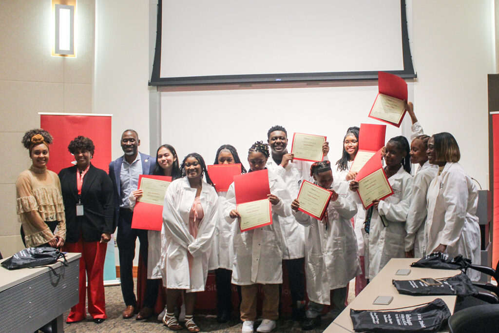 Three leaders stand beside ten high school students who are wearing their new medical white coats and waving their graduation certificates in the air