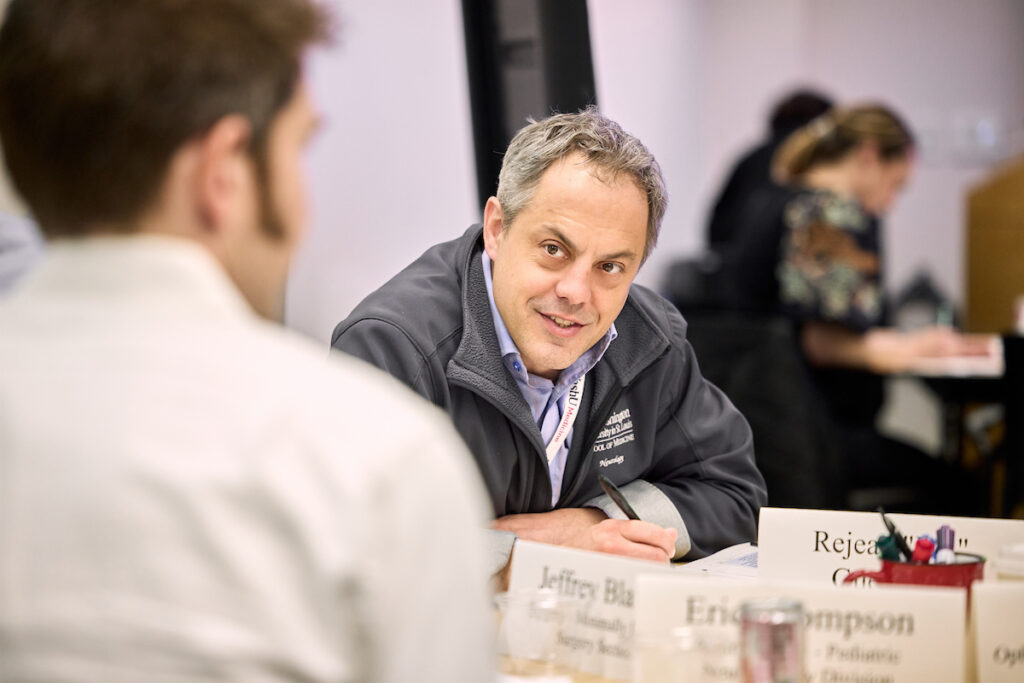 Man sitting at table, wearing a WashU Medicine fleece, listens to a colleague who is wearing a white shirt.