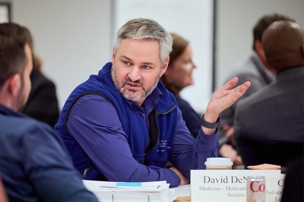 Man sitting at table, wearing a blue fleece, leads a discussion with a colleague who is wearing a blue shirt.