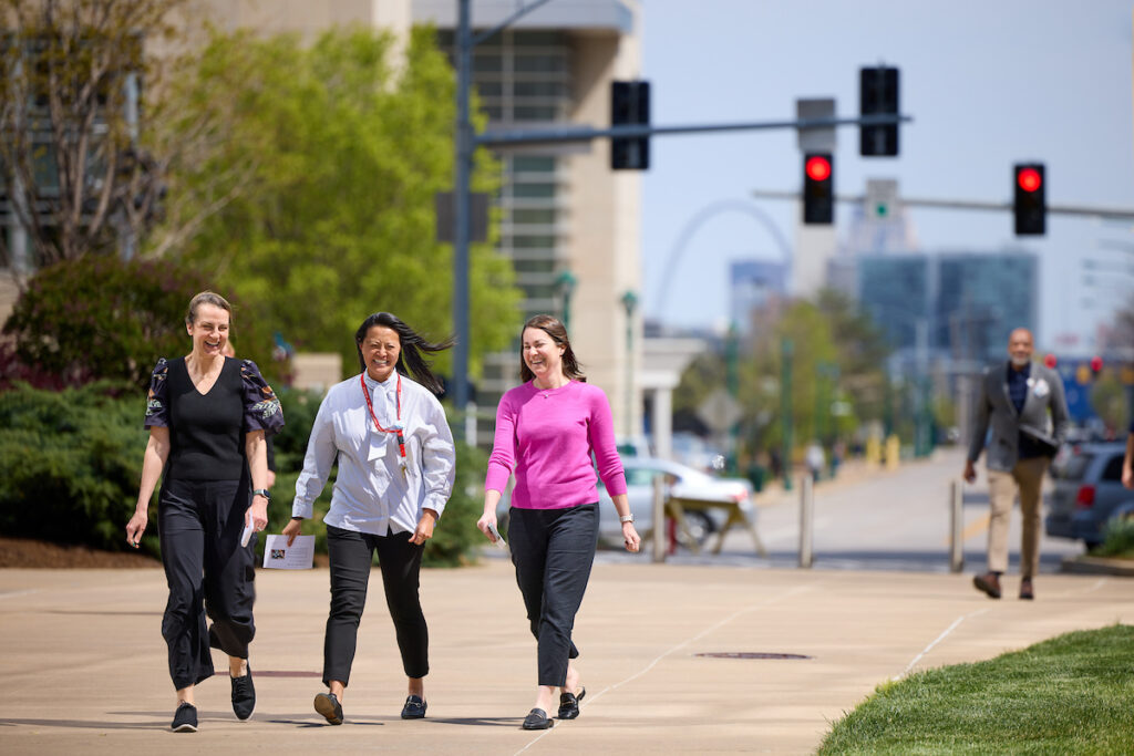 Three women are smiling as they walk together on the medical campus. The St. Louis arch can be seen in the background.