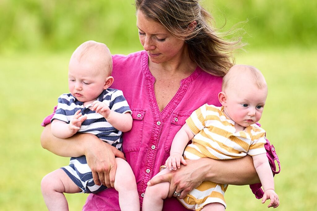 A person in a pink shirt holds two babies outdoors in a grassy field.