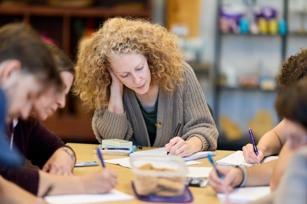 A person with curly hair sits at a table, writing in a notebook while others around them also write, suggesting a collaborative learning or creative session.