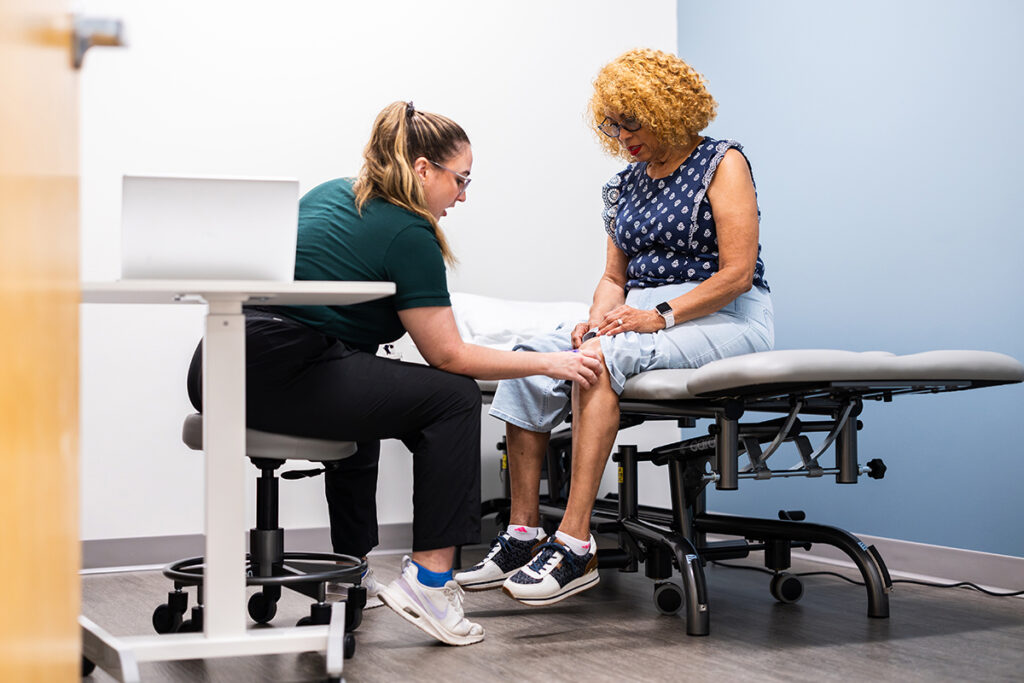 Woman sits on exam table (right) while woman seated to her left touches her knee