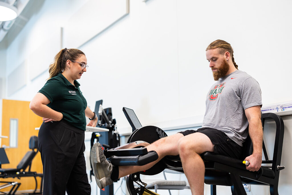Man on right extends his leg while using exercise machine; woman to his left watches and provides guidance