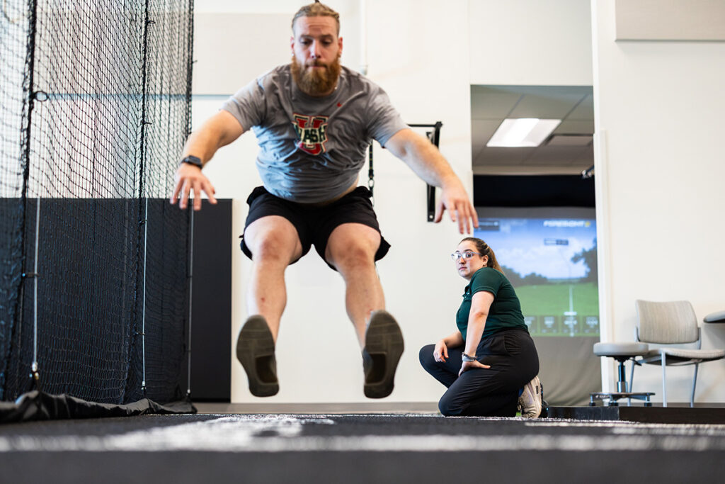 Man jumping toward camera, legs in front of him, while woman in foreground watches his form