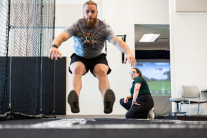Man jumping toward camera, legs in front of him, while woman in foreground watches his form