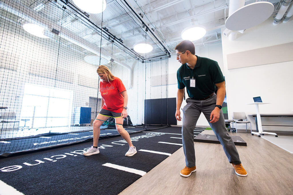 Woman in red exercises with rubber band wrapped around her legs, near her knees. To her right, man in green watches