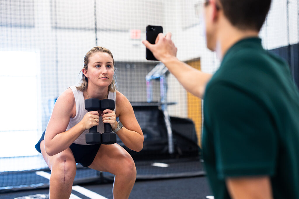 Woman holding dumbbells while in a squat looks at device held by man in green shirt