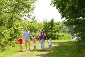 A family of five walks along a sunlit grassy path bordered by lush green trees.