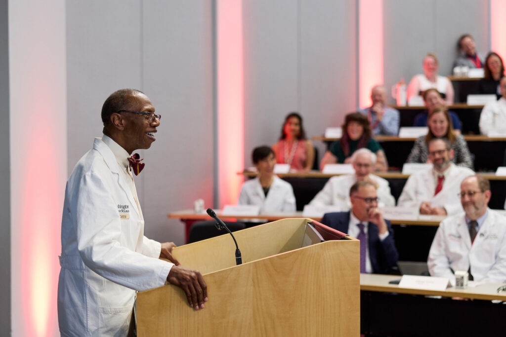 A person in a white lab coat stands at a podium speaking to an audience seated in rows.