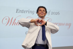 A person in a white coat and blue tie makes a heart shape with their hands while standing in front of a screen displaying "Entering Class of 2026 White Ceremony."