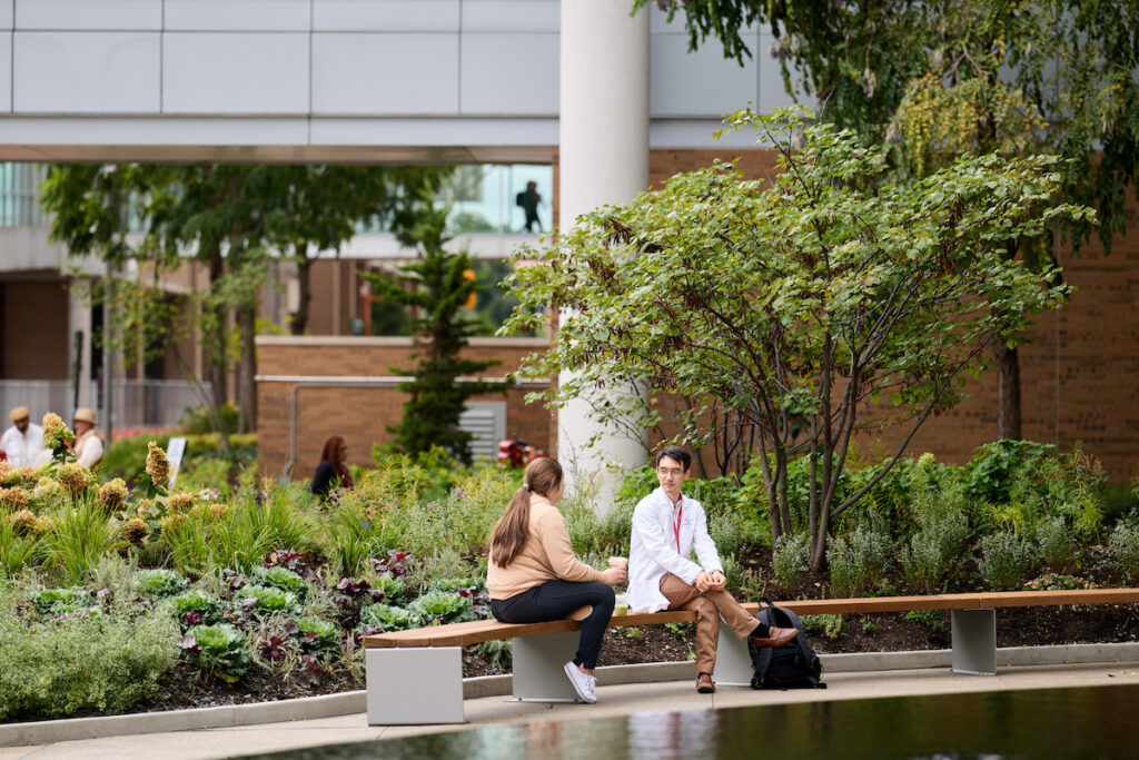 Two individuals sit on a bench in a modern outdoor courtyard, surrounded by greenery and a reflective pool, with buildings and pedestrians visible in the background.