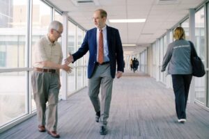 Dean Bruce D. Levy, MD, greets a man while walking through a glass-walled corridor.